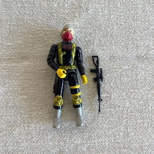 May include: A vintage action figure of a soldier in a black and yellow uniform with a red mask and helmet. The figure has yellow gloves and camouflage knee pads. A black rifle is included.
