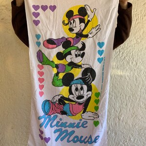 Puede incluir: Toalla de playa blanca con una imagen colorida de tres personajes de Minnie Mouse con ropa de entrenamiento. La toalla tiene el texto "Minnie Mouse" en letras moradas y azules. El fondo es blanco con corazones rosas, morados y verdes.