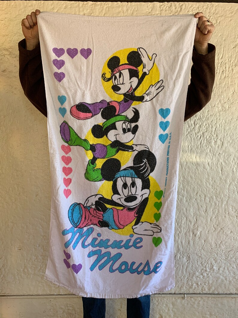 Puede incluir: Toalla de playa blanca con una imagen colorida de tres personajes de Minnie Mouse con ropa de entrenamiento. La toalla tiene el texto "Minnie Mouse" en letras moradas y azules. El fondo es blanco con corazones rosas, morados y verdes.