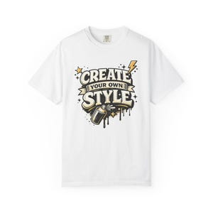 Könnte beinhalten: Weißes T-Shirt mit dem Aufdruck "CREATE YOUR OWN STYLE". Das Design zeigt eine Sprühdose, Sterne und einen Blitz in Schwarz, Gold und Weiß. Ein lässiges Kleidungsstück.