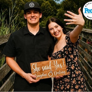 May include: A rectangular wooden sign with the white text She said Yes! and the names Brayden and Destiny separated by a ring icon. The sign is held by two people, one wearing a black shirt and cap, the other a floral dress, showing a ring.