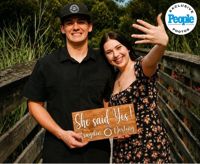 May include: A rectangular wooden sign with the white text She said Yes! and the names Brayden and Destiny separated by a ring icon. The sign is held by two people, one wearing a black shirt and cap, the other a floral dress, showing a ring.