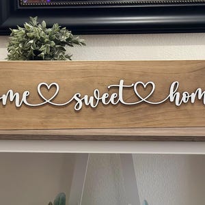 以下が含まれることがあります： 「home sweet home」と白い筆記体で書かれた木製の看板。単語の間に2つのハートがあります。