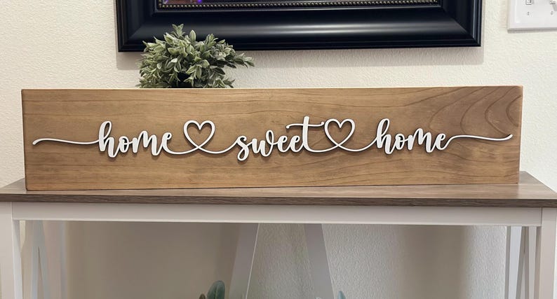 以下が含まれることがあります： 「home sweet home」と白い筆記体で書かれた木製の看板。単語の間に2つのハートがあります。