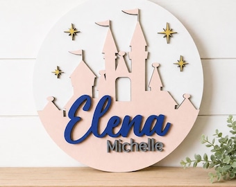 Puede incluir: Un letrero de pared circular de madera blanca con una silueta de castillo en capas de color rosa claro. El nombre Elena está escrito en letra cursiva grande de color azul oscuro, con el nombre Michelle impreso debajo en letras mayúsculas gris oscuro.