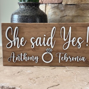 May include: A wooden sign with the text "She said Yes!" in white cursive lettering. Below the text is a white ring with a diamond and the names "Anthony" and "Febronia" in white cursive lettering.