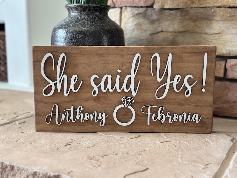 May include: A wooden sign with the text "She said Yes!" in white cursive lettering. Below the text is a white ring with a diamond and the names "Anthony" and "Febronia" in white cursive lettering.