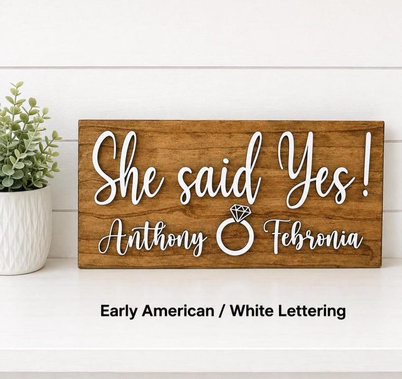 May include: A rectangular wooden sign with a medium brown finish featuring white raised lettering that reads She said Yes! Anthony and Febronia with a white diamond ring icon centered between the names. The text Early American / White Lettering is below.