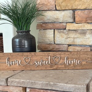 以下が含まれることがあります： 「home sweet home」と cursive で書かれた木製の看板で、各単語の間にハートマークがあります。看板は茶色で、素朴な仕上げになっています。石の表面に置かれています。