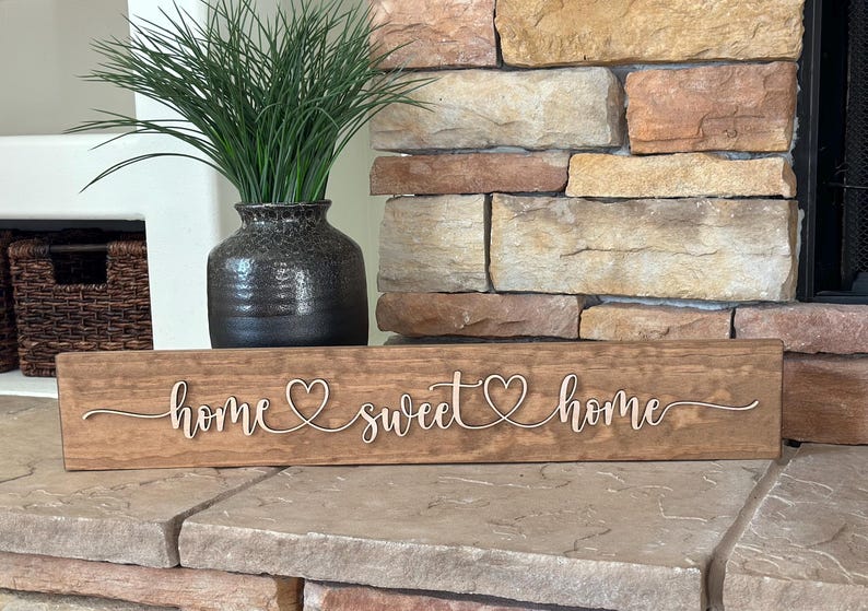 以下が含まれることがあります： 「home sweet home」と cursive で書かれた木製の看板で、各単語の間にハートマークがあります。看板は茶色で、素朴な仕上げになっています。石の表面に置かれています。