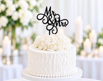 May include: A black cake topper featuring the text Mr & Mrs in a cursive script font with decorative swirls. The topper is positioned on top of a white cake decorated with a cluster of white roses and a beaded border along the base of the cake.
