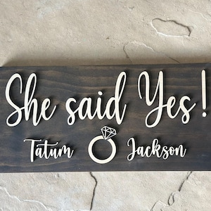 May include: A dark brown wooden sign with the text "She said Yes!" in white lettering. The sign also includes the names "Tatum" and "Jackson" in white lettering, along with a white diamond ring icon.