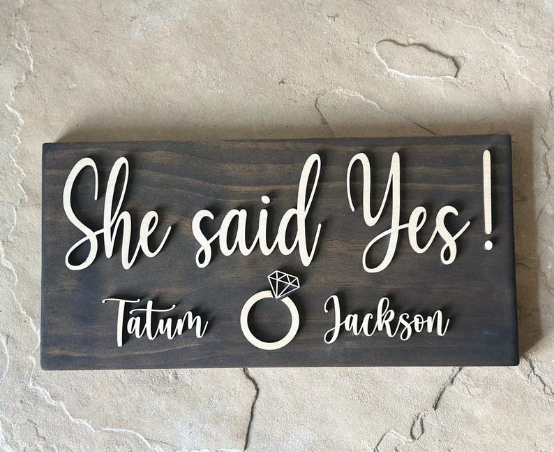 May include: A dark brown wooden sign with the text "She said Yes!" in white lettering. The sign also includes the names "Tatum" and "Jackson" in white lettering, along with a white diamond ring icon.
