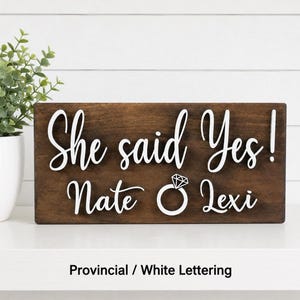 May include: A rectangular dark brown wooden sign featuring white raised lettering that reads "She said Yes!" above the names "Nate" and "Lexi" with a white graphic of a diamond engagement ring positioned between the two names.