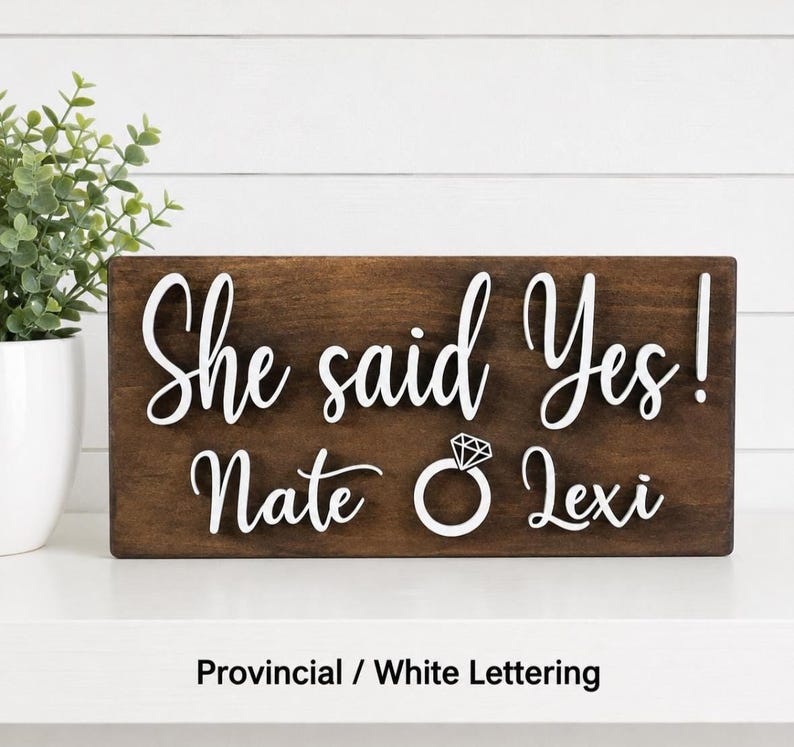 May include: A rectangular dark brown wooden sign featuring white raised lettering that reads "She said Yes!" above the names "Nate" and "Lexi" with a white graphic of a diamond engagement ring positioned between the two names.