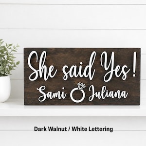 May include: A rectangular dark walnut wooden sign featuring raised white lettering that reads "She said Yes!" above the names "Sami" and "Juliana" separated by a white graphic icon of a diamond engagement ring.