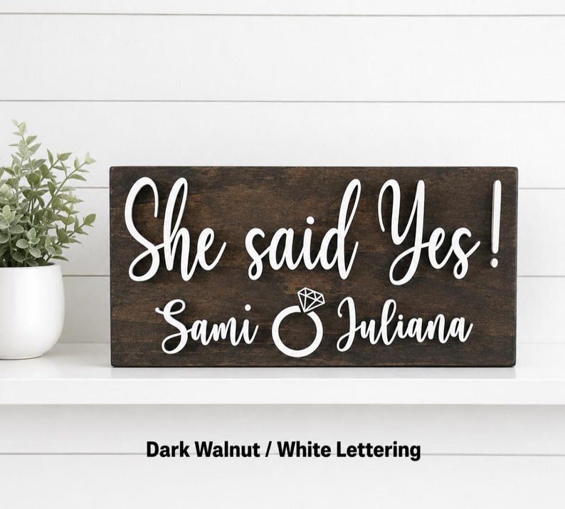 May include: A rectangular dark walnut wooden sign featuring raised white lettering that reads "She said Yes!" above the names "Sami" and "Juliana" separated by a white graphic icon of a diamond engagement ring.