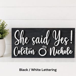 May include: A rectangular black wooden sign featuring raised white lettering that reads She said Yes! Coleton and Nichole, with a white graphic of a diamond ring positioned between the two names. The sign has a smooth, dark finish.