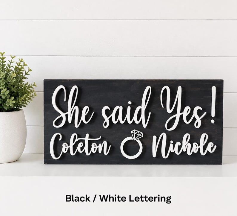 May include: A rectangular black wooden sign featuring raised white lettering that reads She said Yes! Coleton and Nichole, with a white graphic of a diamond ring positioned between the two names. The sign has a smooth, dark finish.