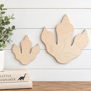 May include: Two light-colored wooden dinosaur footprint cutouts in different sizes. The smooth, natural wood grain is visible on both shapes, which feature three distinct toes and a rounded heel, displayed as flat wall decor items.
