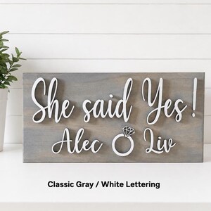 May include: A rectangular gray wooden sign featuring raised white lettering that reads "She said Yes!" above the names "Alec" and "Liv" with a white diamond ring icon positioned between the names. The sign has a smooth, matte finish.