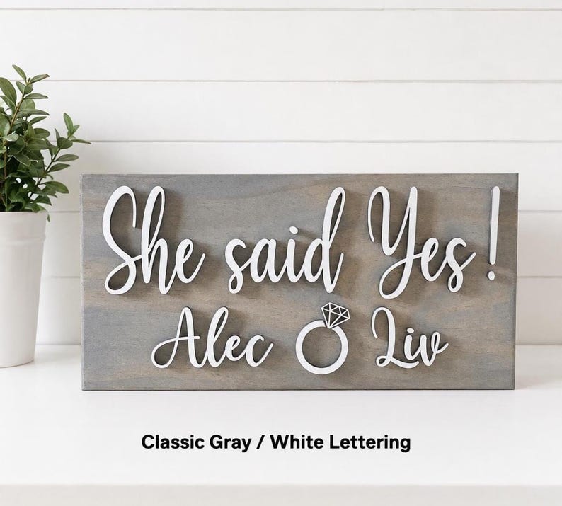 May include: A rectangular gray wooden sign featuring raised white lettering that reads "She said Yes!" above the names "Alec" and "Liv" with a white diamond ring icon positioned between the names. The sign has a smooth, matte finish.