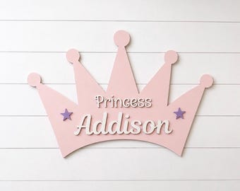 May include: A light pink crown-shaped wall sign featuring the word Princess in white script above the name Addison in larger white script. Two small purple stars are positioned on either side of the name Addison on the crown.