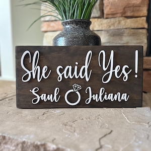 May include: A wooden sign with the text "She said Yes! Saul & Juliana" in white lettering. A diamond ring icon is between the names.