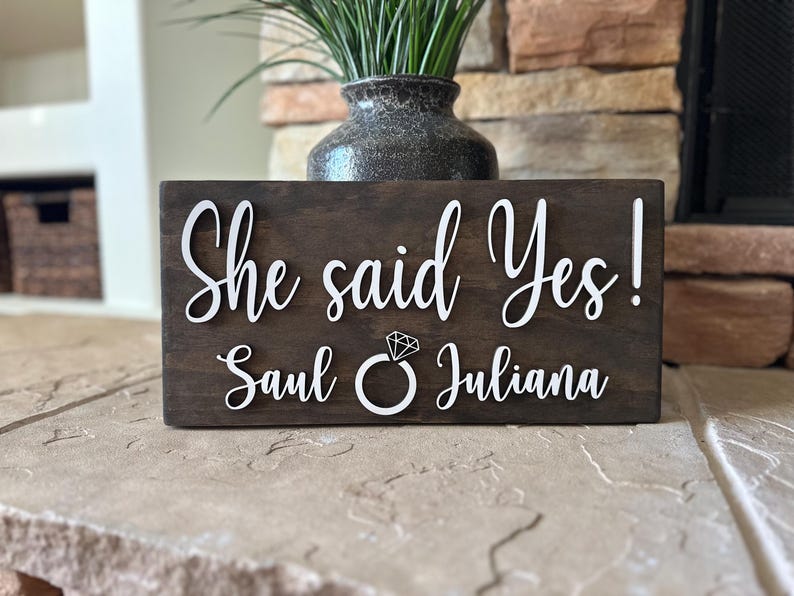 May include: A wooden sign with the text "She said Yes! Saul & Juliana" in white lettering. A diamond ring icon is between the names.