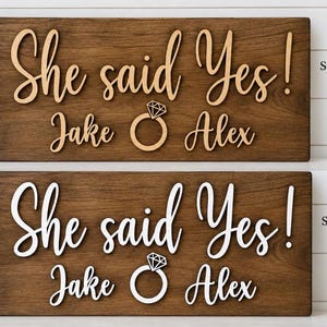 May include: Two rectangular dark walnut wood signs featuring the text She said Yes! with the names Jake and Alex and a diamond ring icon. The top sign has natural wood lettering, while the bottom sign features white lettering on the wood surface.
