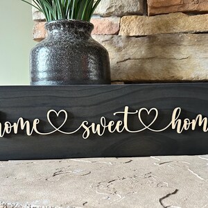 以下が含まれることがあります： 「home sweet home」と cursive で書かれた黒い木製の看板。各単語の間にハートマークがあります。