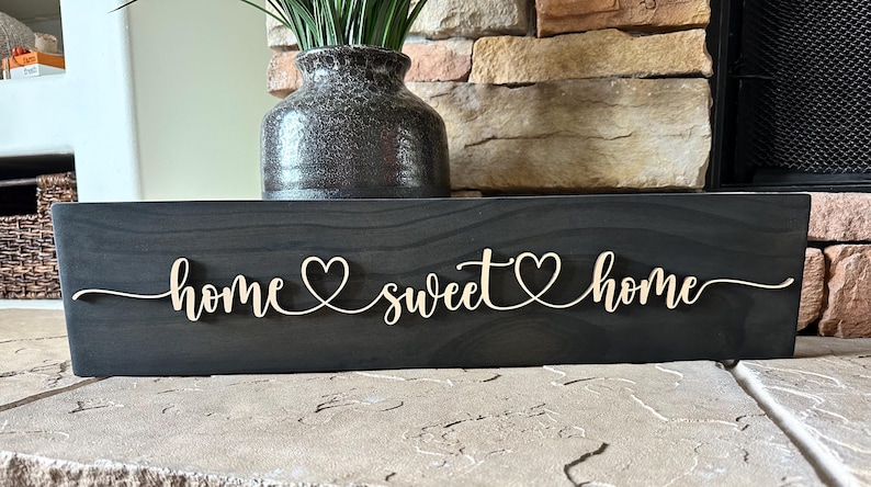 以下が含まれることがあります： 「home sweet home」と cursive で書かれた黒い木製の看板。各単語の間にハートマークがあります。