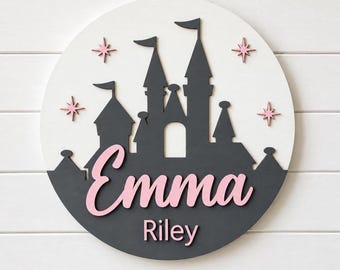May include: A circular wall sign featuring a white background with a dark gray silhouette of a castle. The name Emma is written in large, light pink script, and the name Riley is written below in smaller, light pink block letters with four pink stars.