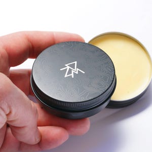 May include: A black tin with a white mountain logo and a topographic pattern on the lid. The tin is open and contains a yellow balm.