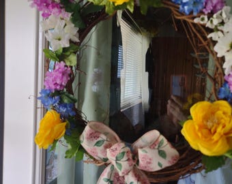 May include: A wreath made of grapevine with a pink floral ribbon. The wreath is decorated with yellow, pink, and blue artificial flowers.