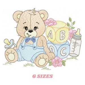 Könnte beinhalten: Stickerei mit einem braunen Teddybär, der eine blaue Latzhose und eine blaue Schleife trägt, mit einem Schnuller sitzt, neben einem Stapel ABC-Blöcken und einer Babyflasche.
