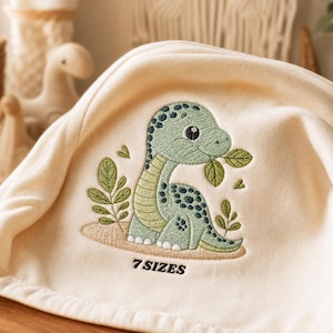 Puede incluir: Una tela de color crema con un diseño bordado de un dinosaurio verde claro con manchas verde oscuro, rodeado de pequeñas hojas y plantas verdes. El texto 7 SIZES está bordado con hilo negro debajo de la ilustración del dinosaurio.
