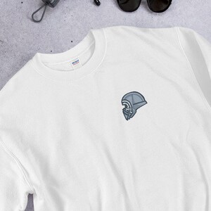 Może przedstawiać: Biała bluza z małym haftowanym wzorem hełmu na lewej piersi.