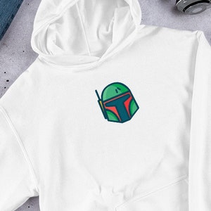 以下が含まれることがあります： 前面に緑と赤のボバ・フェットのキャラクターのグラフィックが描かれた白いフード付きスウェットシャツです。