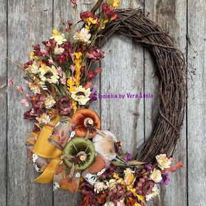 May include: A fall-themed wreath with a brown grapevine base, decorated with a variety of dried flowers in shades of red, yellow, and white. The wreath also features a yellow and brown bow, and three velvet pumpkins in shades of orange, green, and white.