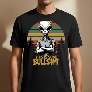 Puede incluir: Camiseta negra con un diseño de alienígena de dibujos animados con los brazos cruzados, sobre un fondo de puesta de sol. El texto amarillo dice "THIS IS SOME BULLSH*T". La camiseta es de cuello redondo.