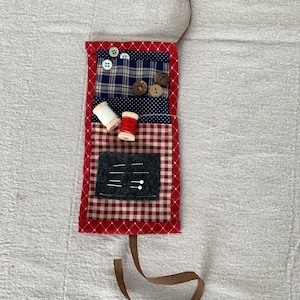 Peut inclure: Un étui à aiguilles en tissu patchwork rouge et bleu avec un ruban brun. L'étui a une poche pour les aiguilles, une poche pour les bobines de fil et une poche pour les boutons.