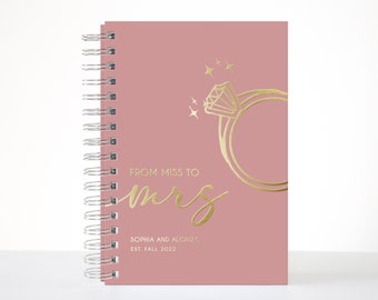 May include: A dusty rose spiral-bound notebook featuring a gold foil illustration of a diamond ring and the text FROM MISS TO mrs, SOPHIA AND ALONZO, and EST. FALL 2022 printed on the front cover in a combination of serif and script fonts.