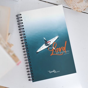 Puede incluir: Un cuaderno azul verdoso con un kayak blanco en una masa de agua. El texto "The Lord will guide you" está escrito en naranja con la referencia bíblica "Isaiah 58:11". El texto "live humbly inspired" está escrito en negro en la parte inferior del cuaderno.
