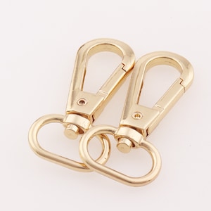 May include: Two gold-toned metal swivel clasps with a D-ring on each. The clasps are open and ready to be attached to a bag or other item.