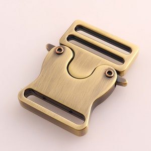 May include: A bronze-colored metal buckle with a unique design. The buckle has a rectangular frame on one side and a curved, interlocking mechanism on the other. It features two small rivets and is likely used for securing straps or closures.