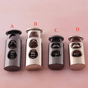 Może przedstawiać: Cztery metalowe blokady sznurka w rzędzie, każda z innym wykończeniem: czarnym, srebrnym i dwoma odcieniami szarości. Każda blokada sznurka ma cylindryczny korpus z dwoma owalnymi otworami i sprężyną w środku.