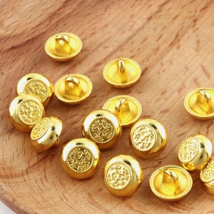 Puede incluir: Un conjunto de 12 botones de color dorado con un diseño decorativo. Los botones son redondos y tienen un pequeño bucle en la parte posterior para coser.