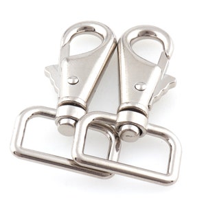 May include: Two silver-colored swivel snap hooks with rectangular D-rings. The hooks have a spring-loaded clasp and are designed for attaching to straps or other hardware. The metal has a shiny, reflective surface.