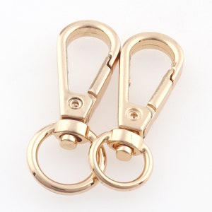 Könnte beinhalten: Zwei goldfarbene Metall-Drehverschlüsse mit einem Ring an jedem Ende. Die Verschlüsse sind offen und bereit, an einer Tasche oder einem anderen Gegenstand befestigt zu werden.